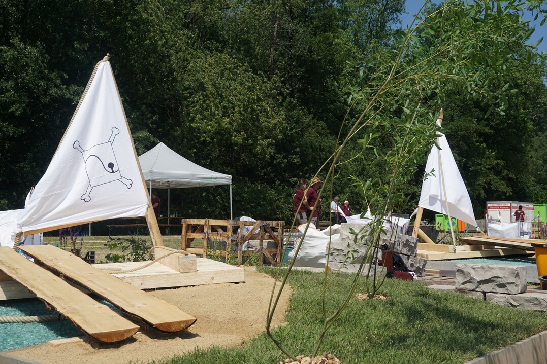 Die Bauaufgabe war das Herzstck des Berufswettkampfes der Landschaftsgrter:innen. Innerhalb von zehn Stunden mussten alle elf Teams einen identischen Garten nach Plan bauen. Dieser stand ganz im Zeichen des Inselparks und bildete neben Ruinenteilen der Wilhelmsburg auch ein angreifendes Piraten-Flo mit Gangway und eine stilisierte Fluss-Szenerie aus blauem Glasbruch und blau blhenden Pflanzen ab.