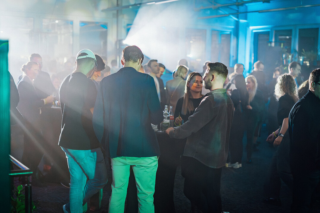 Gäste der Firmeneinweihung stehen in lockerer Runde zusammen und unterhalten sich in stimmungsvoll beleuchtetem Ambiente.