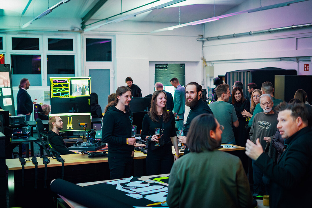 Gäste der Firmeneinweihung im Entwicklungsbereich von hTRIUS im Gespräch, umgeben von Arbeitsplätzen und Technik.